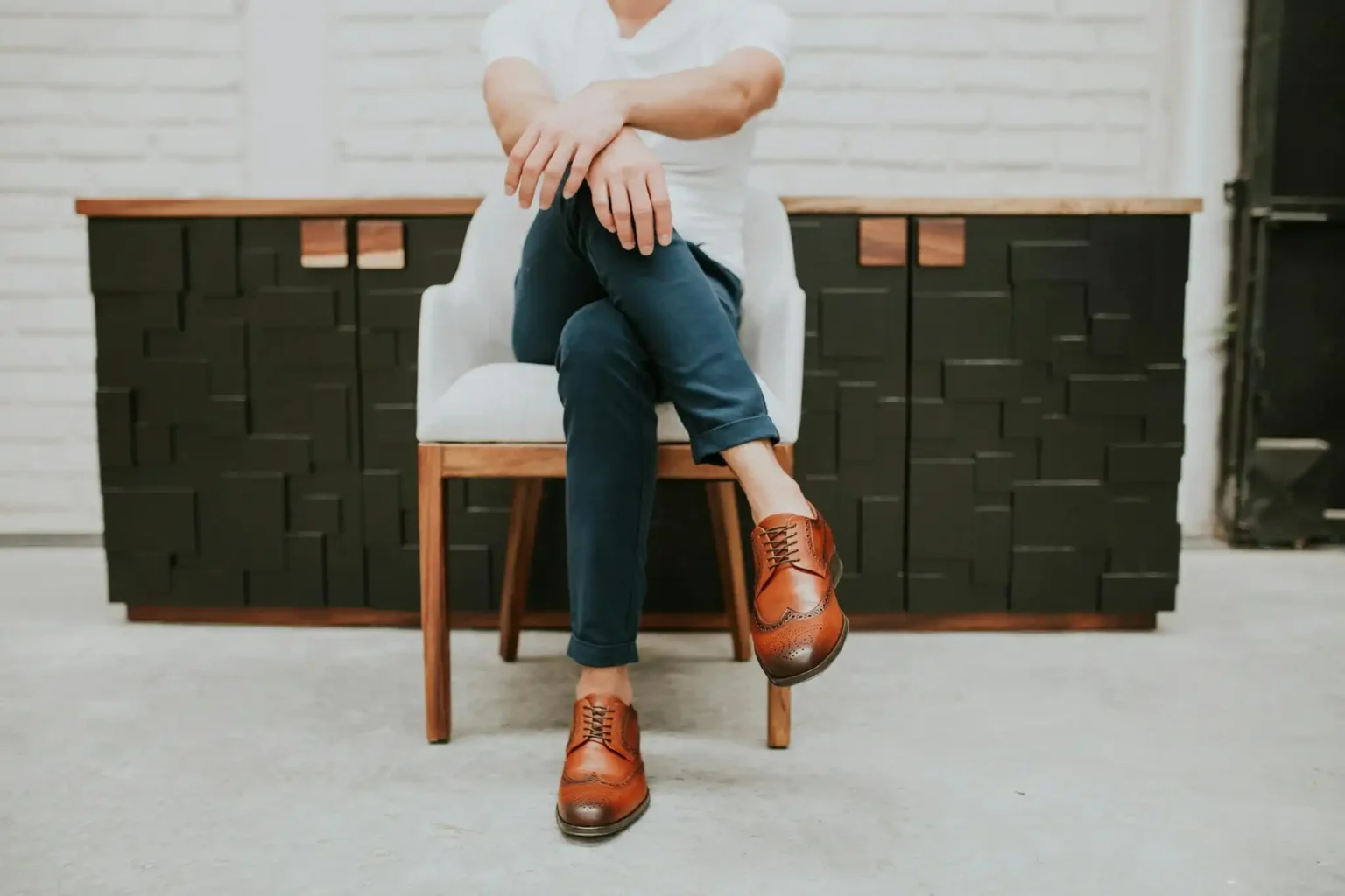 Homme élégant en t-shirt blanc et chaussures en cuir, jambes croisées, assis dans un intérieur moderne